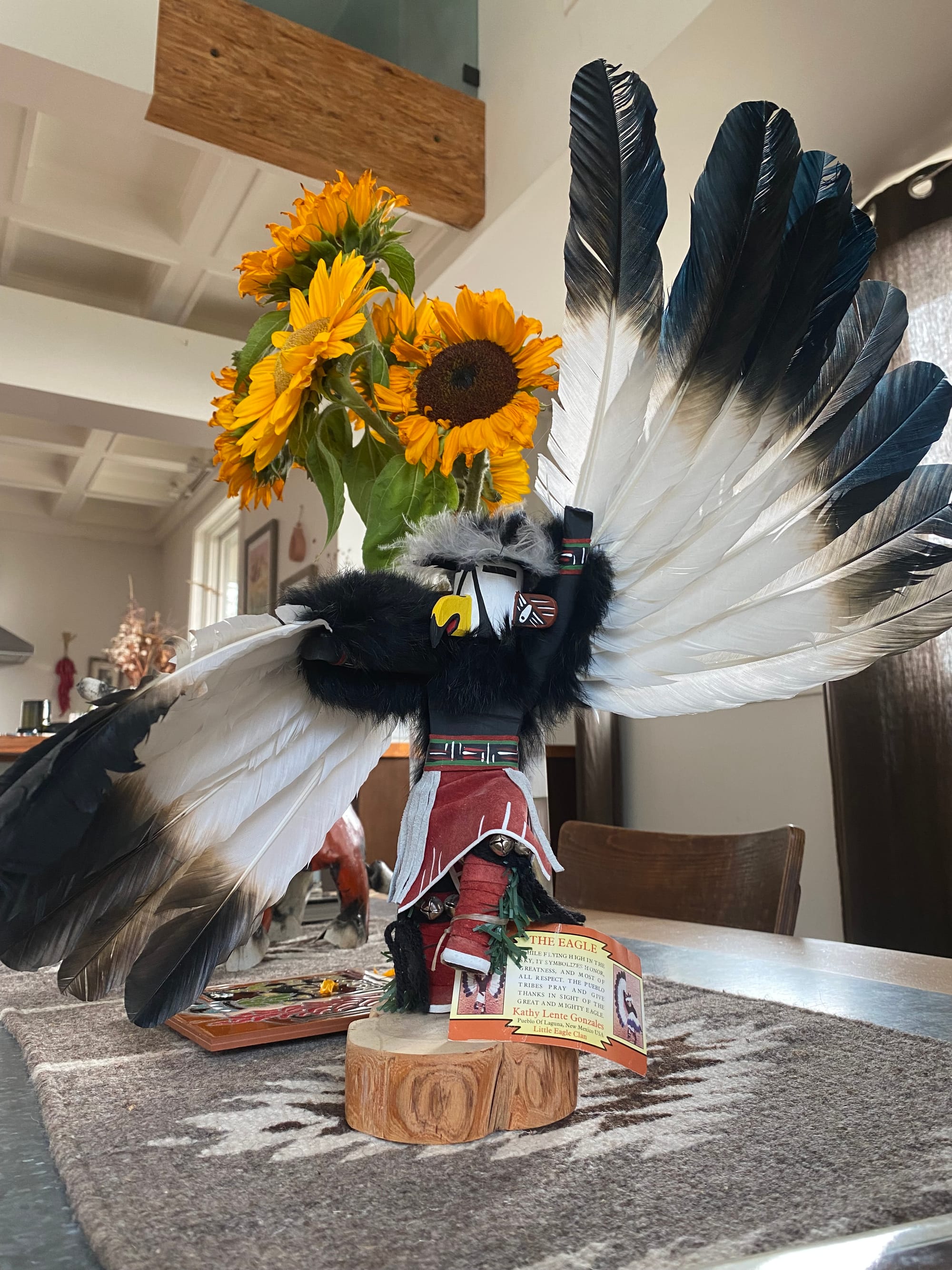 Photo of a kachina doll with feathered wings outstretched with sunflowers in a vase behind on a grey Navajo  table runner 