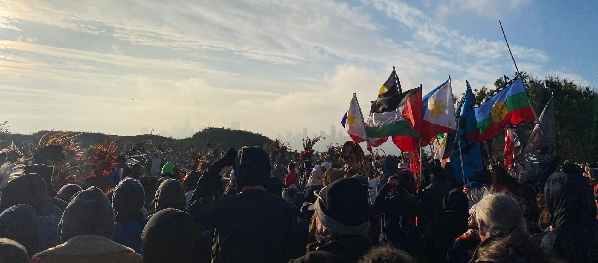Sun rises over a crowd of people watching native ceremony with a cityscape in the background. 